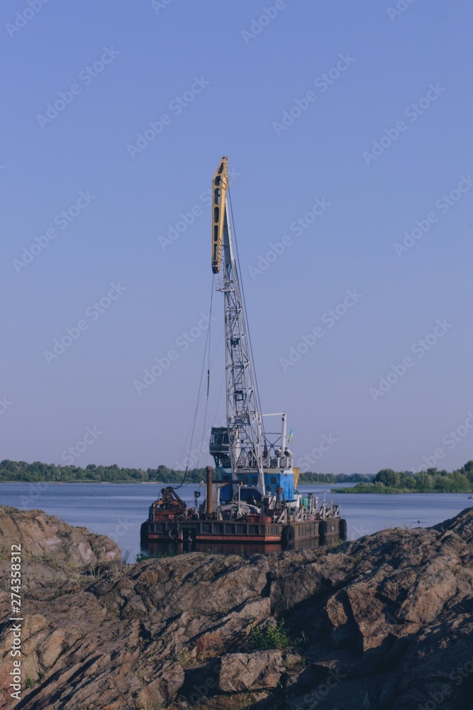 River landscape with boat crane on the water. Blue sky without clouds. The rocks on the foreground. River landscape with boat crane on the water. Blue sky without clouds. The rocks on the foreground.