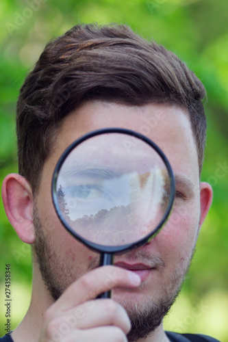 Close-up photo of a man in the magnifying glass. Handsome young man holding magnifying glass close to face, big eyes and serious expression.