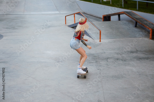 Young lady with blonde hair, with tattooed arms, wears in a red T-shirt and denim shorts, with a knitted bandana on her head, enjoying longboarding in skate park.