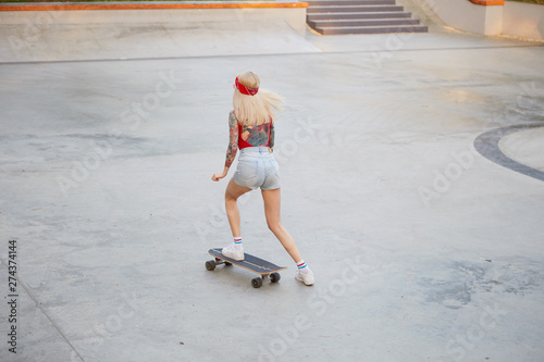 Young tattooed lady with blonde hair raiding on skateboard, wears in a red T-shirt and denim shorts, with a knitted bandana on her head, enjoying the day in skate park.