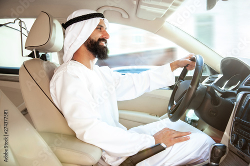 Successful arab saudi businessman riding his car or driving full of emotions at sunny day. Male model as an enterpreneur. Concept of business, finance, modern technologies, start up, economy.