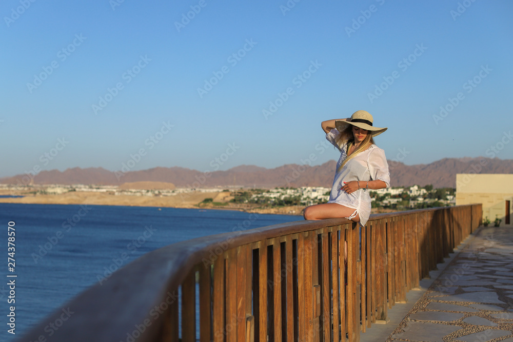 Portrait of a Sexy beautiful tanned woman relaxing in swimsuit and with light beach dress on terrace with blue sea and sky on background. Summer Vacation Concept.