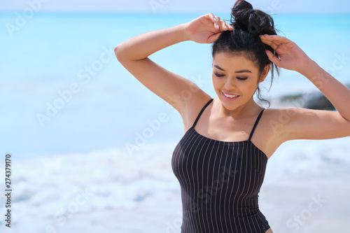 Beautiful latin woman in black bikini on tropical beach. Portrait of happy young woman smiling at sea. Brunette tanned girl in swimwear enjoying and walking on beach.