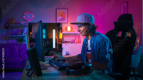 Professional Gamer Playing First-Person Shooter Online Video Game on His Powerful Personal Computer with Colorful Neon Led Lights. Young Man is Wearing a Cap. Living Room Lit with Pink Lamps. Evening. photo