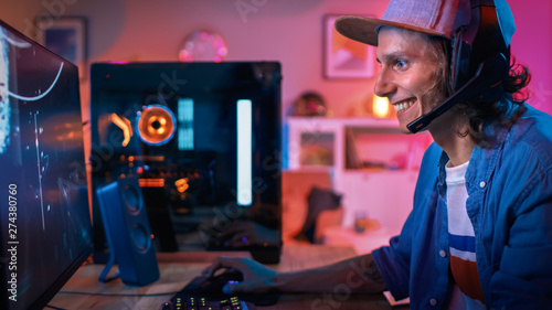 Gamer Puts His Headset with a Mic On and Starts Playing Shooter Online Video Game on His Personal Computer. Room and PC have Colorful Neon Led Lights. Young Man is Wearing a Cap. Cozy Evening at Home.