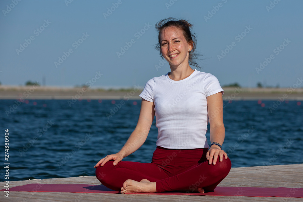 Fototapeta premium Young girl practices yoga on the shore of the lake, the concept of enjoying privacy and concentration, sunlight