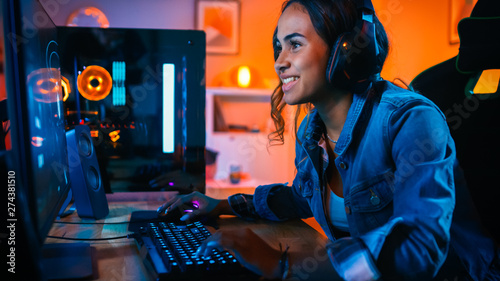 Pretty and Excited Black Gamer Girl in Headphones is Playing First-Person Shooter Online Video Game on Her Computer. Room and PC have Colorful Neon Led Lights. Cozy Evening at Home. photo
