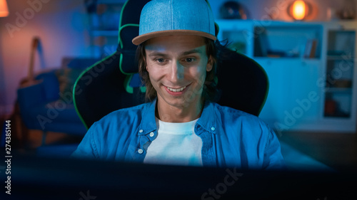 Handsome Surprised and Excited Young Man Watching an Action Video on a Computer. He is Happy and Smiling. Screen Adds Reflections to His Face. Cozy Room is Lit with Warm Light.