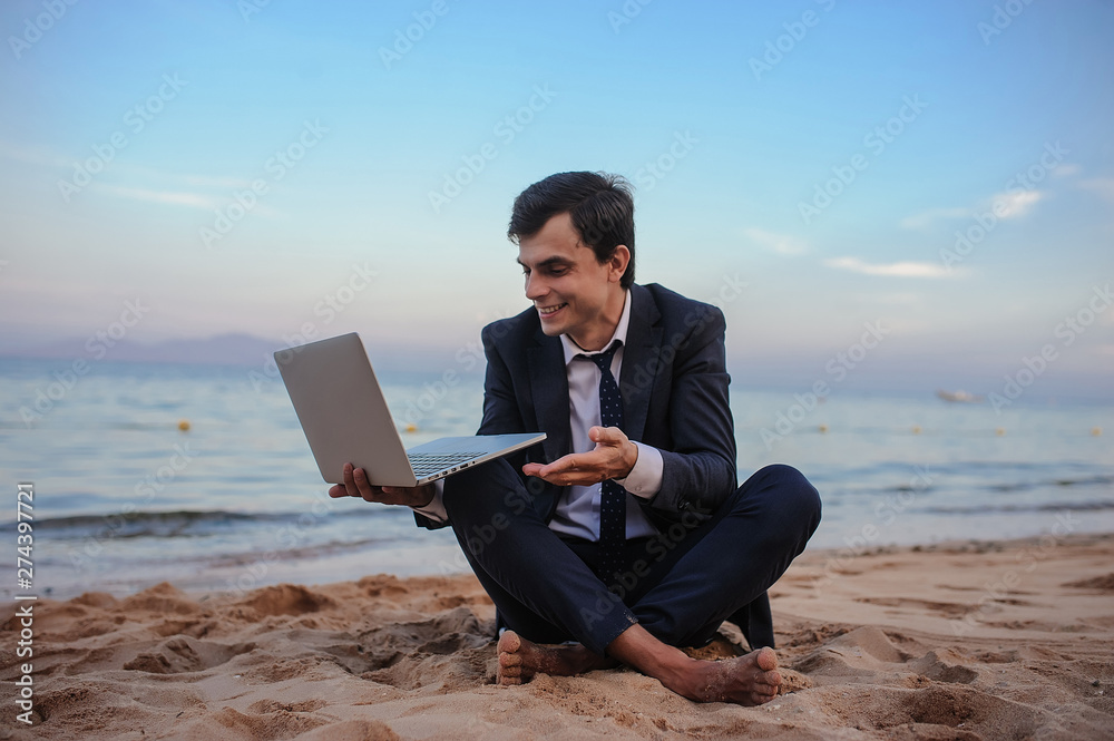 close up photo of a young man in suit with laptop working on the beach and talking to someone