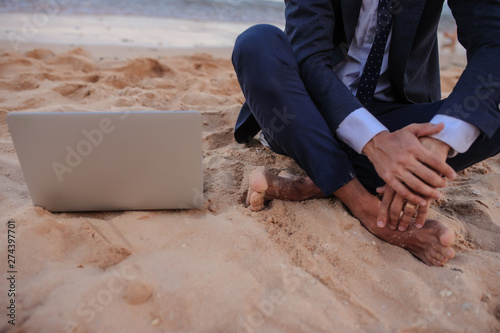 lateral view of a young man in suit with laptop sitting on the beach working on the seaside photo