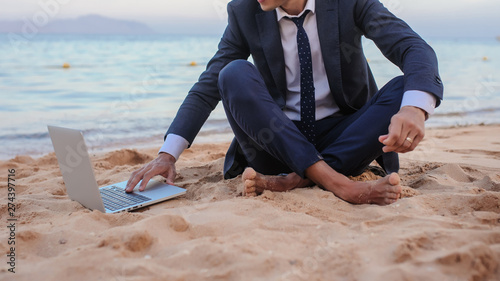 close up photo of a young man in suit with laptop working on the beach and talking to someone