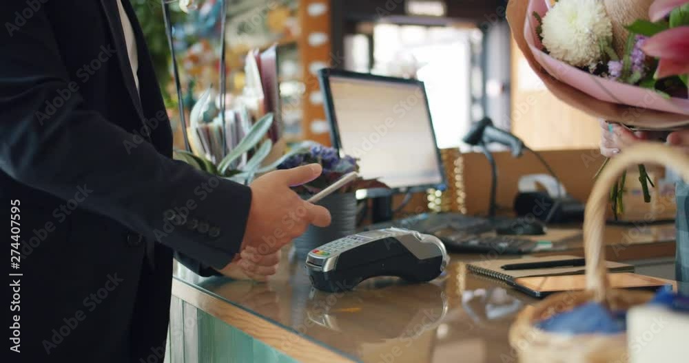 Businessman in suit is buying bouquet of flowers paying online with smartphone holding device making payment in florist's shop. People and gadgets concept. Businessman in suit is buying bouquet of flowers paying online with smartphone holding device making payment in florist's shop. People and gadgets concept.