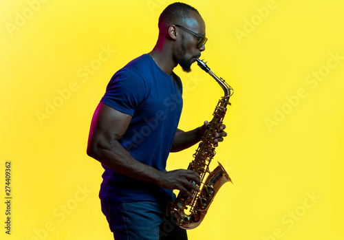 Canvas Print Young african-american jazz musician playing the saxophone on yellow studio background in trendy neon light