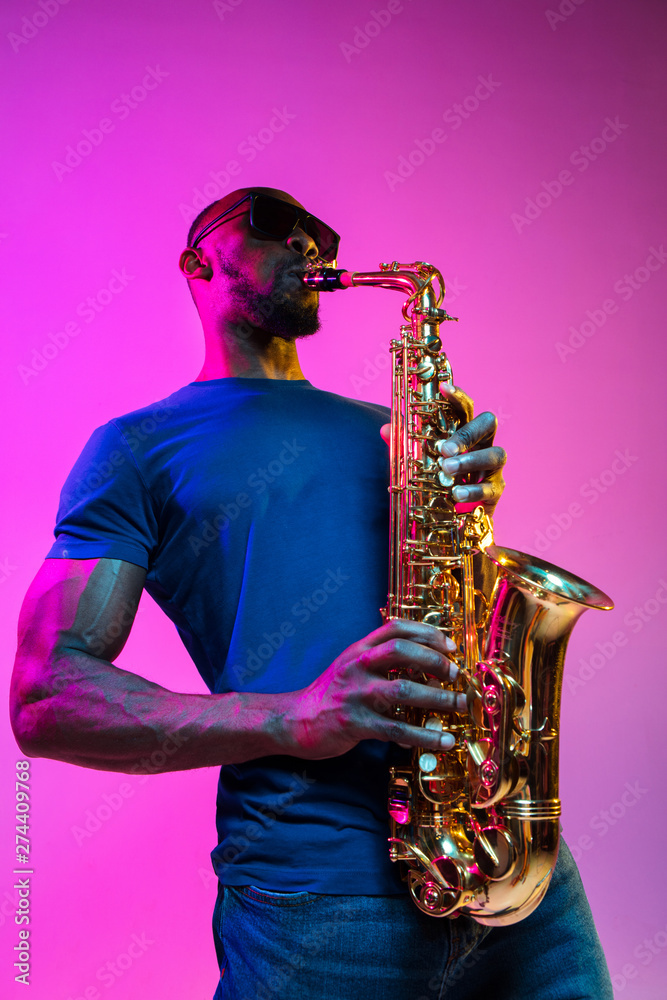 Fototapeta premium Young african-american jazz musician playing the saxophone on pink studio background in trendy neon light. Concept of music, hobby. Joyful attractive guy improvising. Colorful portrait of artist.