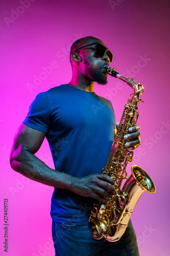 Young african-american jazz musician playing the saxophone on pink studio background in trendy neon light. Concept of music, hobby. Joyful attractive guy improvising. Colorful portrait of artist.