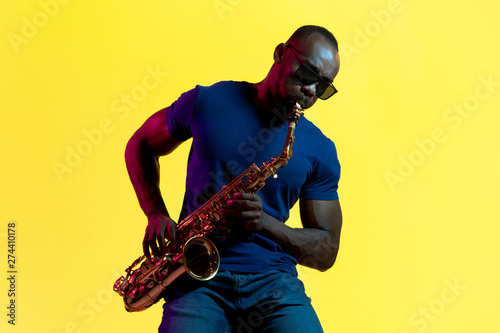 Photography Young african-american jazz musician playing the saxophone on yellow studio background in trendy neon light