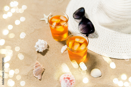vacation  travel and summer concept - two glasses of aperitif cocktails with ice cubes  sun hat  sunglasses and seashells on beach sand