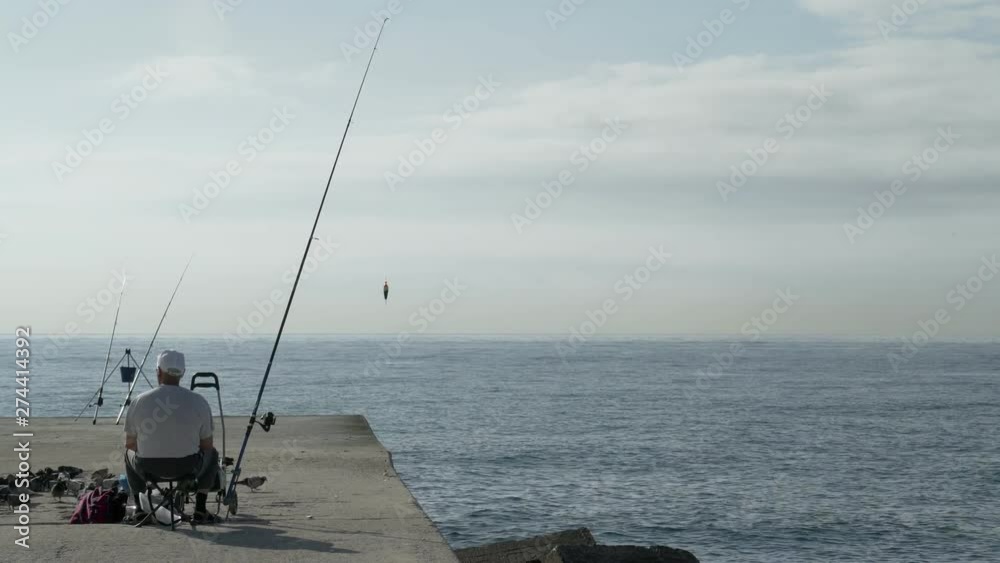 An old man fishing on the pier. Grandpa chilling with a fishing rod at the edge of concrete dock. Summer fishing under a blue sky of Barcelona. Sea and clouds. Panoramic view of the ocean. Harbour. An old man fishing on the pier. Grandpa chilling with a fishing rod at the edge of concrete dock. Summer fishing under a blue sky of Barcelona. Sea and clouds. Panoramic view of the ocean. Harbour.