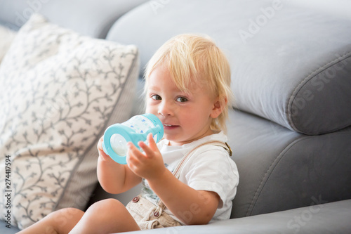 Adorable baby boy drinking milk from a bottle in a white sunny living room.