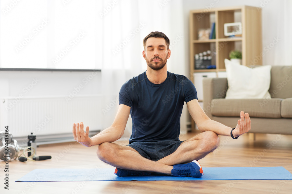 Fototapeta premium sport, fitness and healthy lifestyle concept - man meditating in lotus pose on yoga mat at home