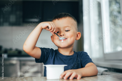 Cute boy in the kitchen eating yogurt from a white container for yogurt, there is a place for advertising