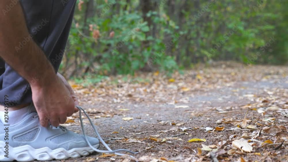 Close up of young sportsman ties shoe in early autumn forest. Athlete tying shoelaces on sneakers before jogging during training outdoor. Feet of strong man jogging along path at nature. Low view Close up of young sportsman ties shoe in early autumn forest. Athlete tying shoelaces on sneakers before jogging during training outdoor. Feet of strong man jogging along path at nature. Low view