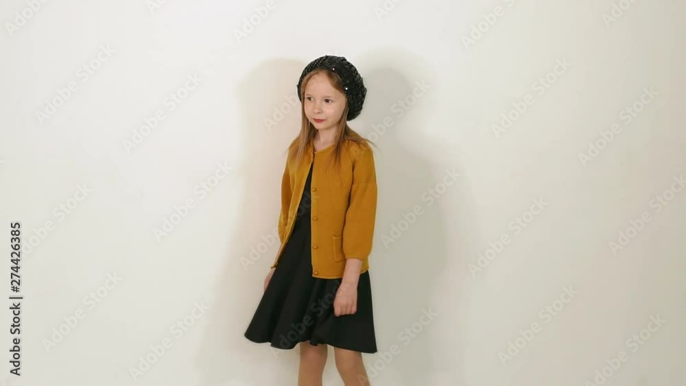 Portrait of cute little girl in black shiny hat, black dress and brown jacket in Studio on white wall background. Slow motion. Portrait of cute little girl in black shiny hat, black dress and brown jacket in Studio on white wall background. Slow motion.