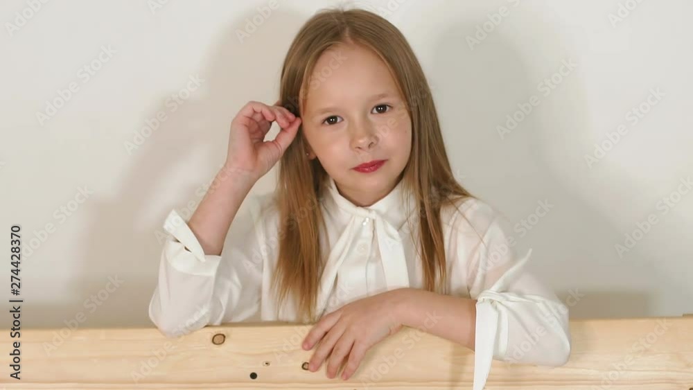 Cute blonde baby girl is standing leaning on a wooden bench in a room with a white wall. Portrait of a little girl near the eco-bench in the Studio with a white background. Slow motion. Cute blonde baby girl is standing leaning on a wooden bench in a room with a white wall. Portrait of a little girl near the eco-bench in the Studio with a white background. Slow motion.