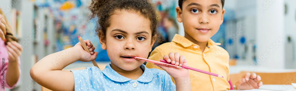 panoramic shot of cute african american kid biting pencil near adorable child panoramic shot of cute african american kid biting pencil near adorable child