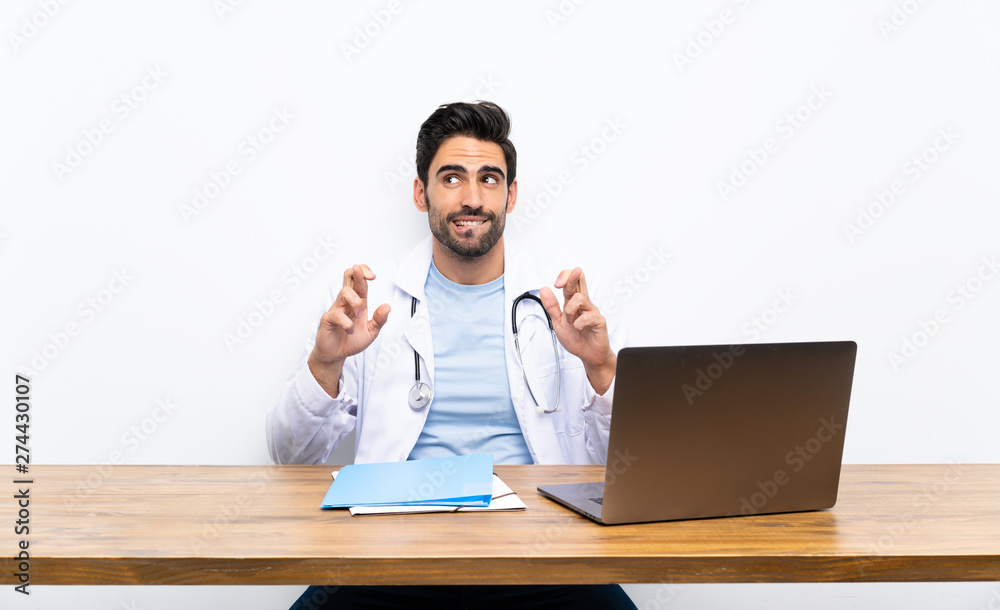 Young doctor man with his laptop over isolated wall with fingers crossing