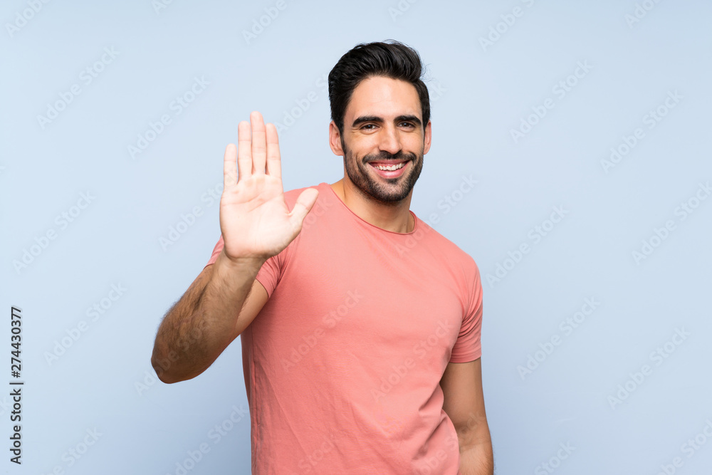 Handsome young man in pink shirt over isolated blue background saluting with hand with happy expression Handsome young man in pink shirt over isolated blue background saluting with hand with happy expression