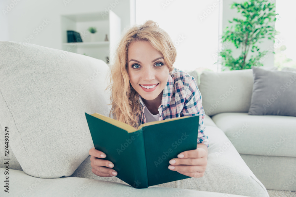 Close-up portrait of nice attractive lovely charming cute cheerful glad nerd wavy-haired girl in checked shirt lying on divan reading new publishing at white light style interior house room indoors Close-up portrait of nice attractive lovely charming cute cheerful glad nerd wavy-haired girl in checked shirt lying on divan reading new publishing at white light style interior house room indoors