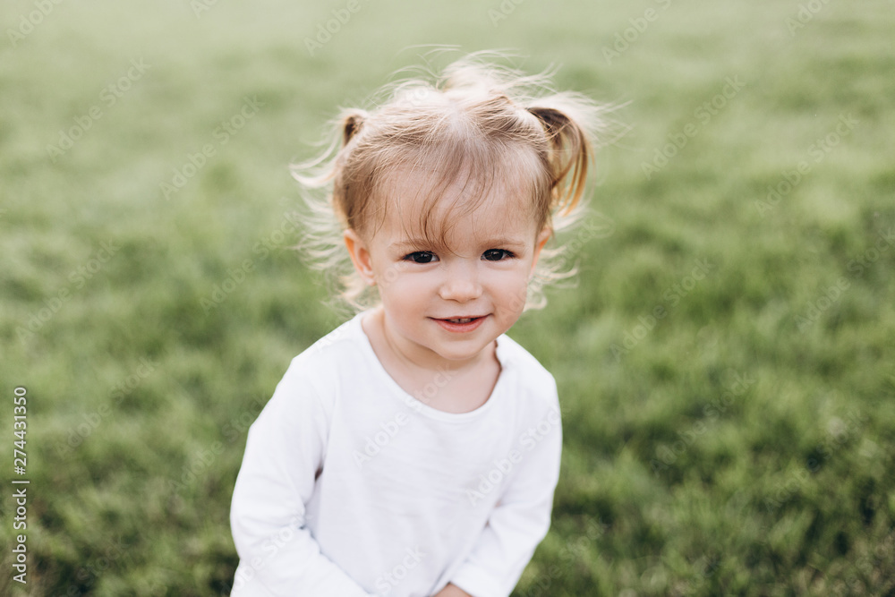 little baby girl with blond hair and two tails, wearing a white T-shirt standing on green grass in nature, smiling little baby girl with blond hair and two tails, wearing a white T-shirt standing on green grass in nature, smiling