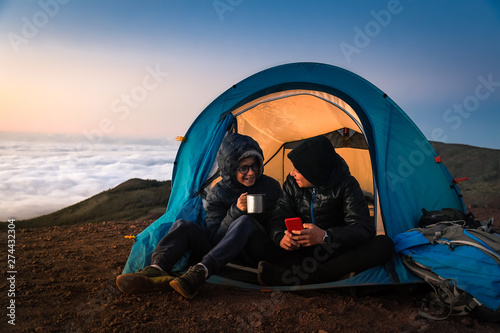 Two boys playing with smartphone sitting in a camping tent. Couple of young tourists enjoying high mountain vacation at sunset. Beautiful panorama in background. Adventure trip and open air concept