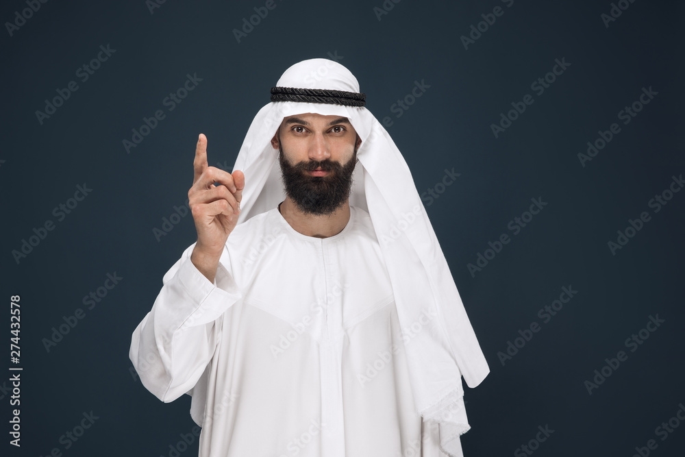 Half-length portrait of arabian saudi businessman on dark blue studio background. Young male model smiling and pointing. Concept of business, finance, facial expression, human emotions. Half-length portrait of arabian saudi businessman on dark blue studio background. Young male model smiling and pointing. Concept of business, finance, facial expression, human emotions.