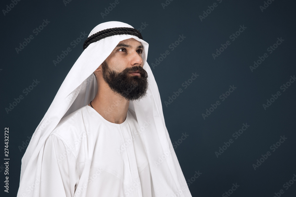 Half-length portrait of arabian saudi businessman on dark blue studio background. Young male model standing and looks thoughtful. Concept of business, finance, facial expression, human emotions.