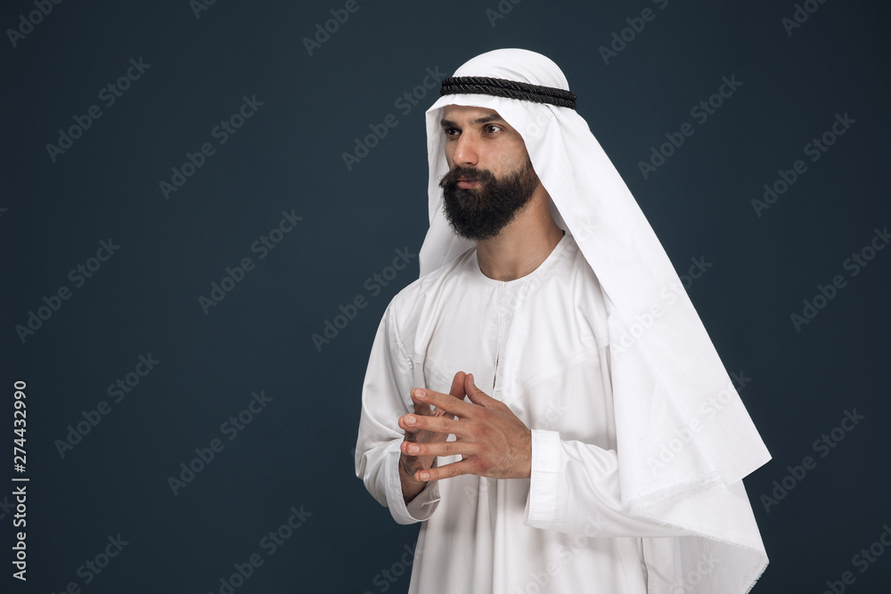 Half-length portrait of arabian saudi businessman on dark blue studio background. Young male model standing and looks thoughtful. Concept of business, finance, facial expression, human emotions. Half-length portrait of arabian saudi businessman on dark blue studio background. Young male model standing and looks thoughtful. Concept of business, finance, facial expression, human emotions.