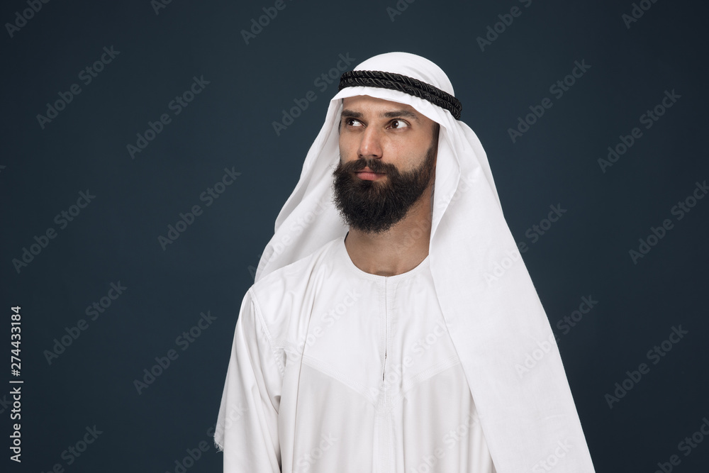 Half-length portrait of arabian saudi businessman on dark blue studio background. Young male model standing and looks thoughtful. Concept of business, finance, facial expression, human emotions.