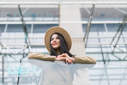 Young cheerful woman in hat and coat standing on balcony and smiling outdoors. Beautiful and fashionable hipster girl in eyeglasses touching hat and posing on city street. Woman in modern city outfit