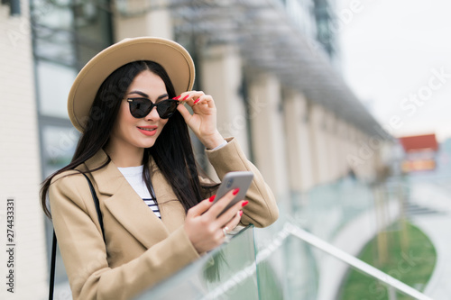 Beautiful girl in a beige coat, holds a smartphone in her hands, stands in front of the building's urban background, looks at the phone screen. Stylish lady uses a smartphone