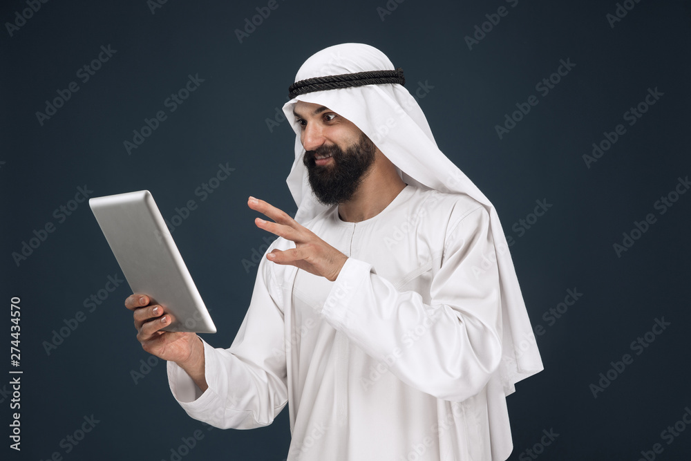 Half-length portrait of arabian saudi businessman on dark blue studio background. Young male model using tablet or gadget. Concept of business, finance, facial expression, human emotions, technologies Half-length portrait of arabian saudi businessman on dark blue studio background. Young male model using tablet or gadget. Concept of business, finance, facial expression, human emotions, technologies