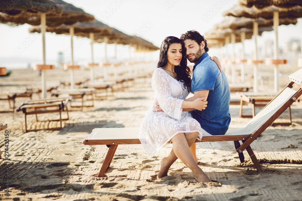 A man and a woman hugging with their eyes closed, sitting on a chair. Couple spending time together at the beach in Rimini, Italy