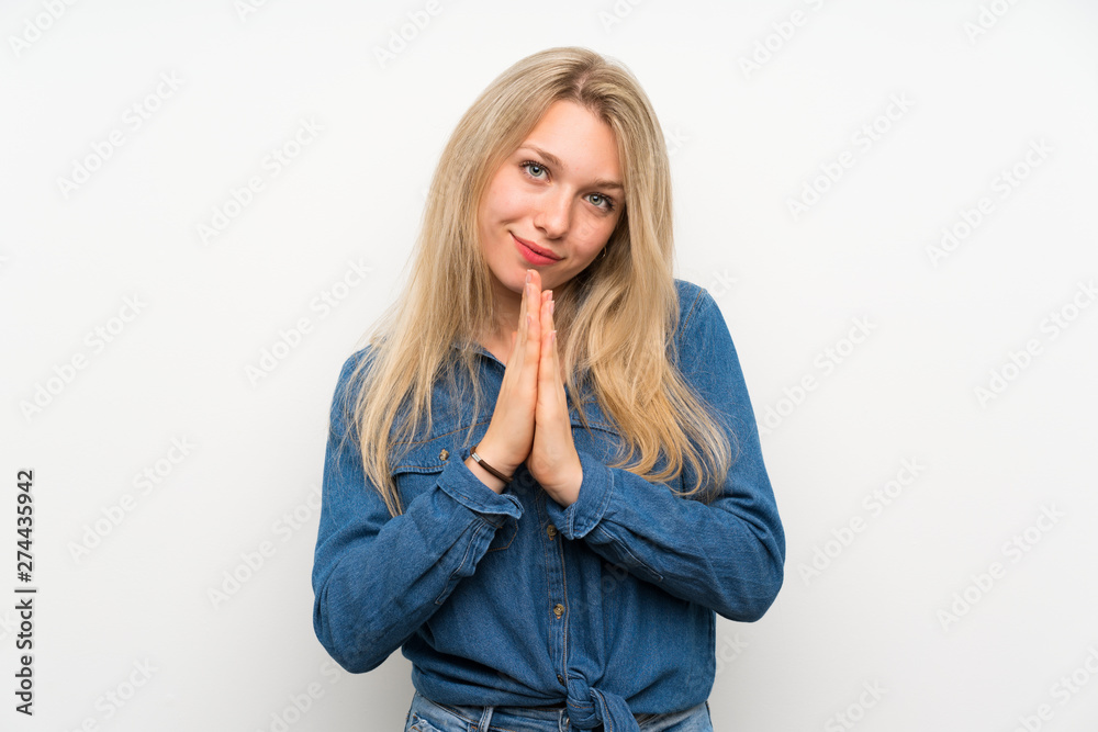 Young blonde woman over isolated white wall keeps palm together. Person asks for something Young blonde woman over isolated white wall keeps palm together. Person asks for something