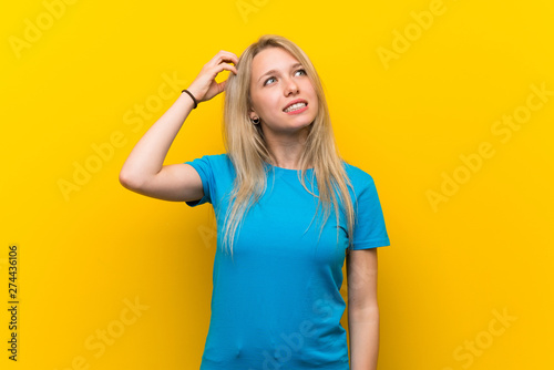 Young blonde woman over isolated yellow background having doubts while scratching head