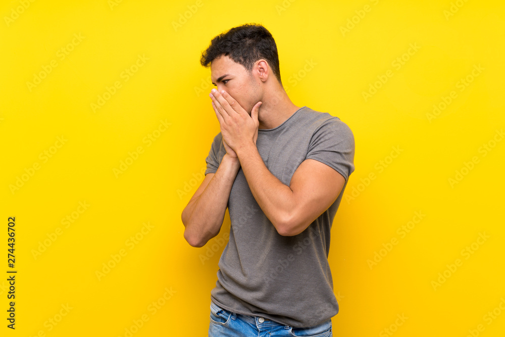 Handsome man over isolated yellow wall covering mouth and looking to the side Handsome man over isolated yellow wall covering mouth and looking to the side