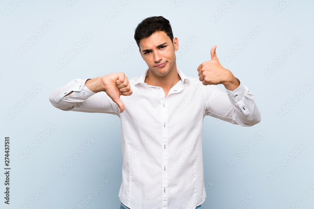 Handsome man over blue wall making good-bad sign. Undecided between yes or not