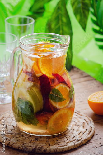 Glass jug of white sparkling wine sangria decorated with citrus slices and season fruits on wooden table. 