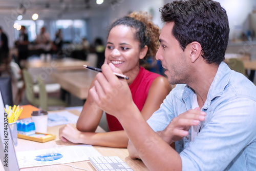 Two business people colleagues talking about planning project in the workplace. Man holding pencil in his hand with woman sitting on desk with computer and documents talking work together in office.