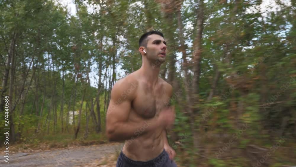 Handsome muscular man with wireless headphones sprinting fast along trail near forest at early autumn. Athletic guy running at nature. Young sportsman training outdoor. Healthy active lifestyle