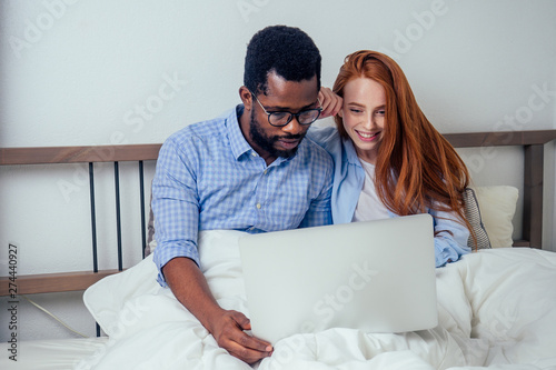 ginger redhaired european female and handsome afro african male together hugging lying in bedroom at home cozy apartment and watching news on laptop.lifestyle interracial family photo